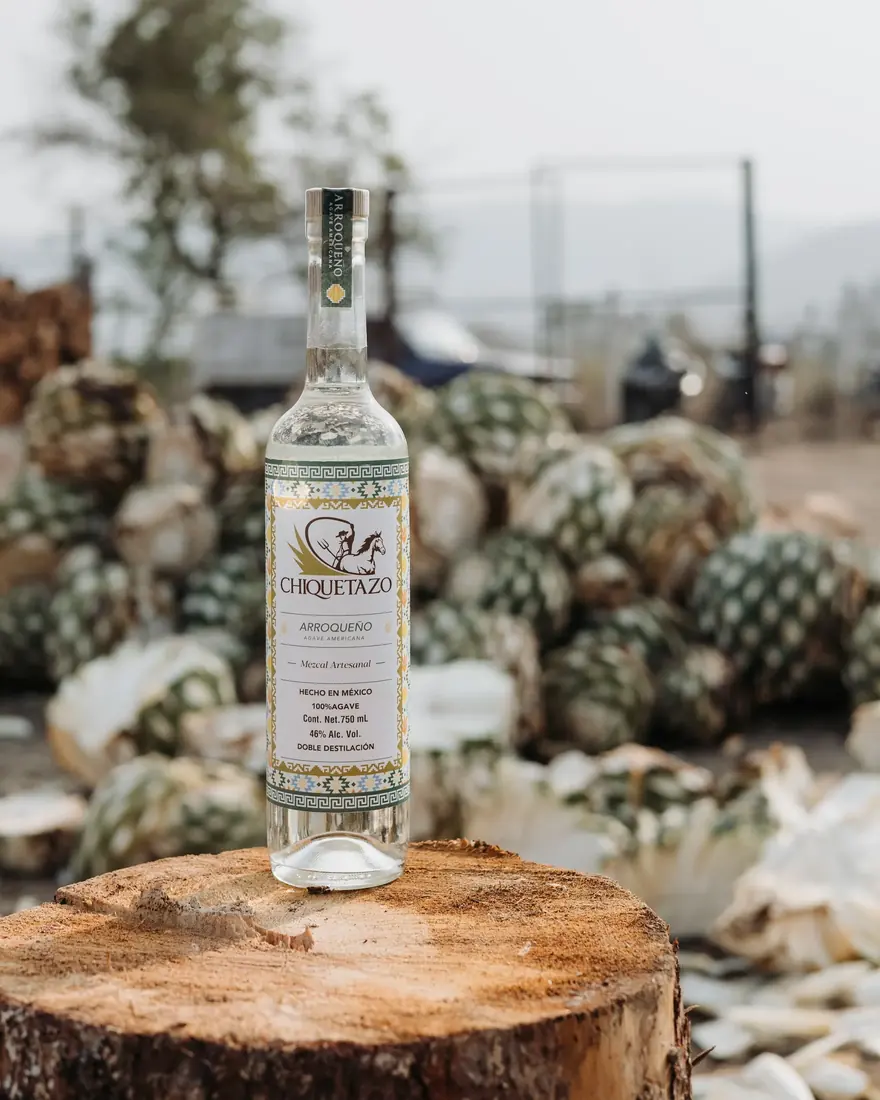 Bottle of Chiquetazo Arroqueño on a tree trunk, with a blurred pile of agave hearts in the background.