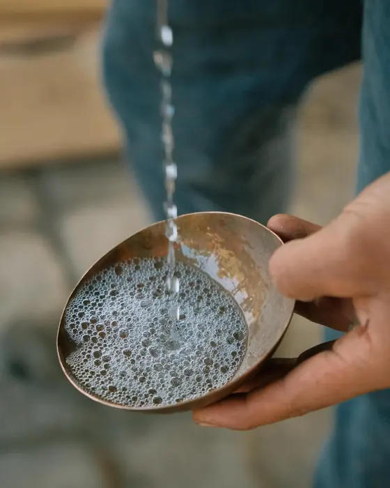 Hands holding a metal cup under the thin stream of mezcal, with the pearls forming on the surface.